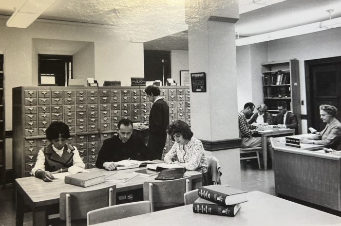Image description: black-and-white photograph of the Missionary Research Library and staff, showing two women and two men in the foreground seated at tables reading books and papers.
