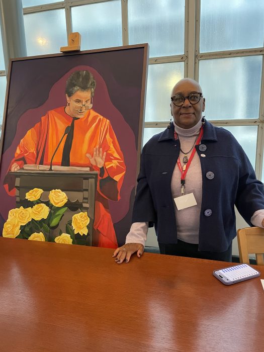 Betty Bolden, a Black woman with glasses and short hair, stands next to a portrait of Dr. Delores Williams, a Black woman in a red robe 