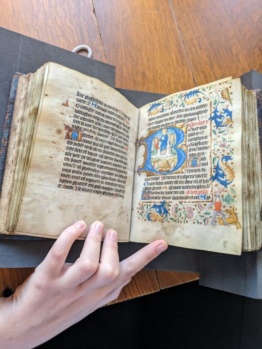 A white hand holds open an illuminated Book of Hours with colorful illustrations