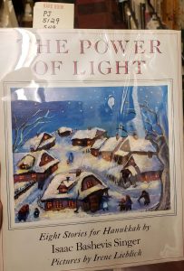 Text reading "The Power of Light: Eight Stories for Hanukkah by Isaac Bashevis Singer." Image of houses covered in snow at night, with people walking around.