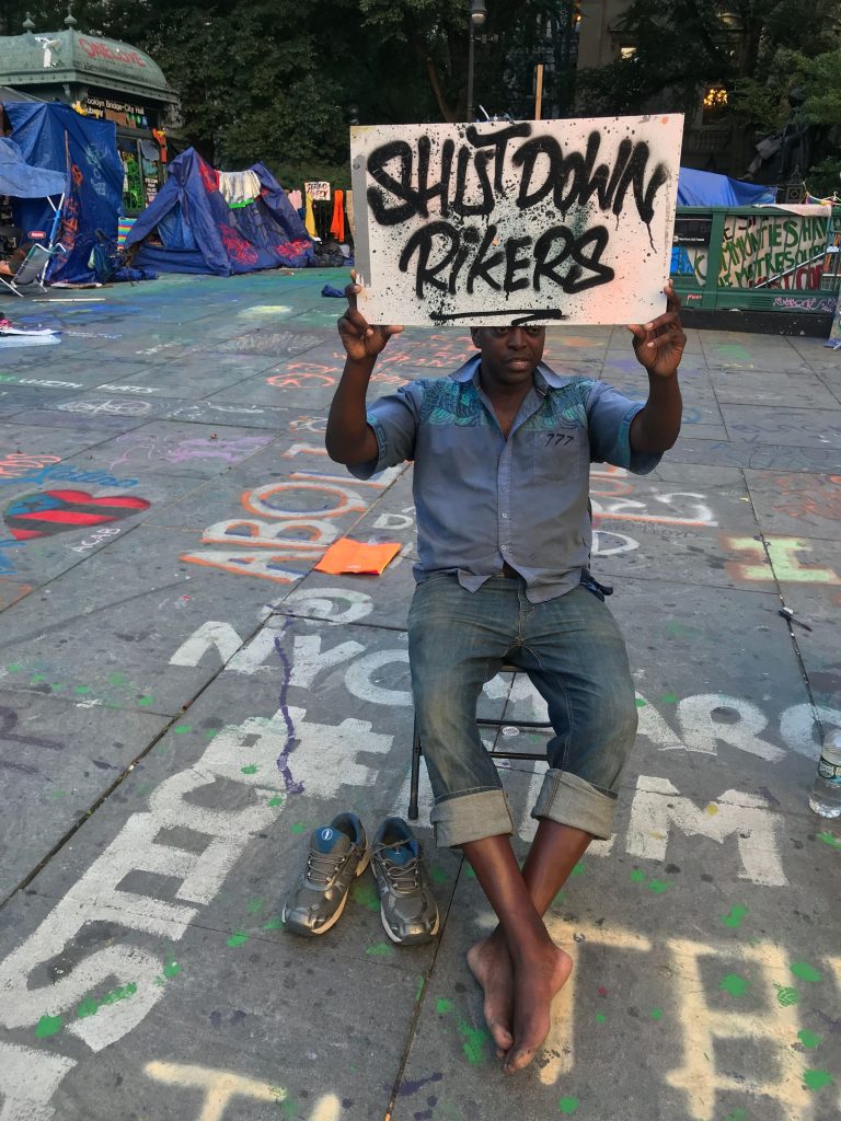seated man in a blue button down shirt and jeans sits in a chair outside a protest encampment holding a sign that says shut down rikers. 