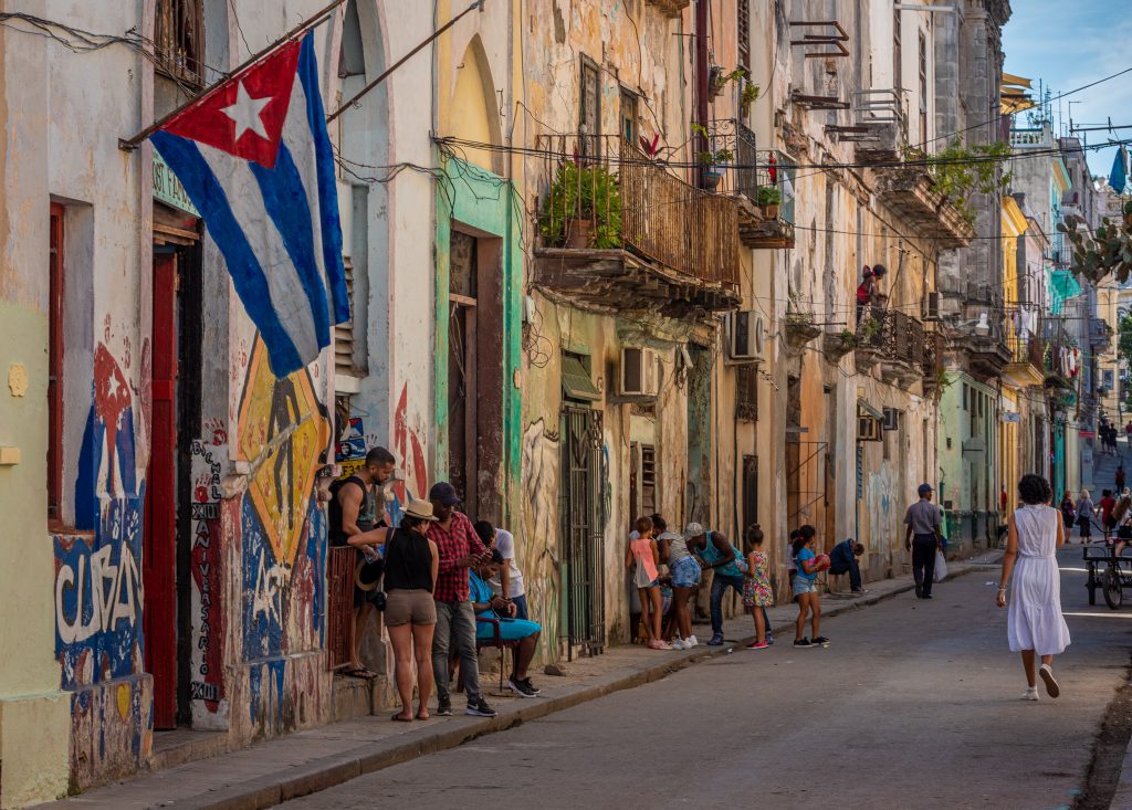 people outside their homes in a Cuban street with wrought iron balconies, faded, but colorfully painted walls, and graffiti signaling Cuban pride, including a Cuban flag hanging from a pole