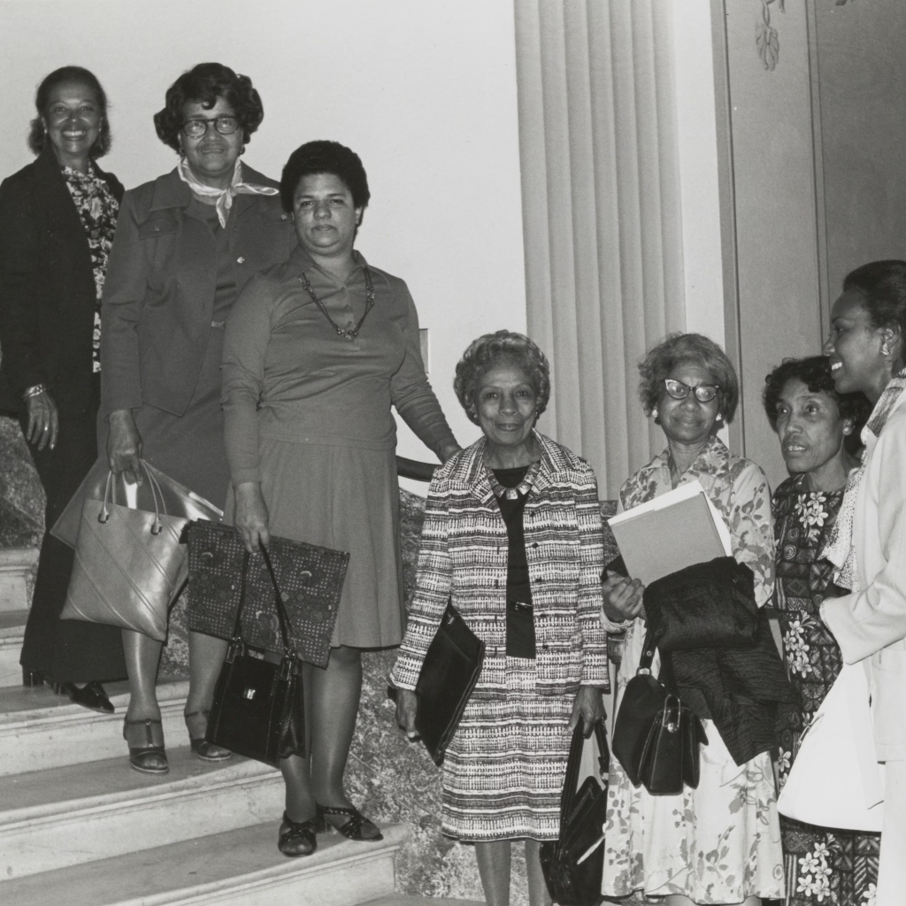 Seven black women dressed up in their 1970s businesswear, lined up on a marble staircase. 